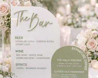 Puede incluir: Un letrero de bar de acrílico blanco con las palabras "The Bar" en escritura verde. Debajo, una lista de bebidas: cerveza, vino y licores. Un letrero más pequeño enumera las bebidas especiales: The Nala-Politan y Clyde's Old Fashioned.