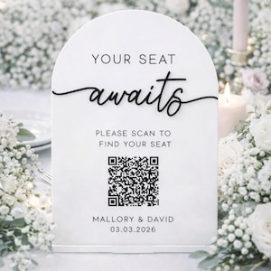 May include: A white wedding sign with an arched top. The sign reads "YOUR SEAT awaits" in black script, with a QR code and the names "Mallory & David" and the date 03.03.2026. The sign is surrounded by white flowers.