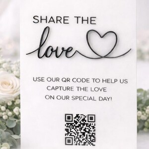 May include: A white acrylic wedding sign with black text. The sign says "SHARE THE love" with a heart symbol. Underneath, it reads "USE OUR QR CODE TO HELP US CAPTURE THE LOVE ON OUR SPECIAL DAY!" A QR code is at the bottom.
