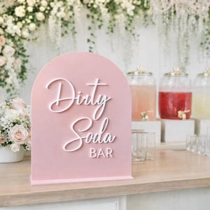 May include: A pink arched sign with the words "Dirty Soda BAR" in white script. The sign is on a wooden table, with floral arrangements and beverage dispensers in the background. Clear glasses are arranged on the table.