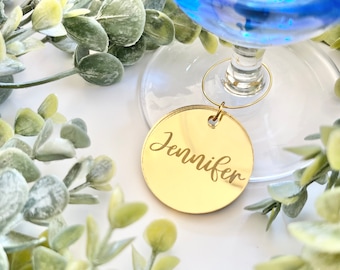 May include: Gold round glass charm with the name Jennifer engraved on it. The charm is attached to a blue glass wine glass.