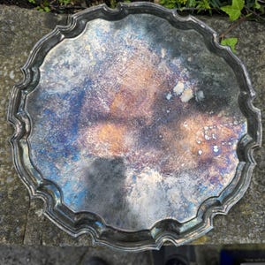 May include: A round silver-toned metal tray with a scalloped, decorative edge. The surface of the tray displays significant oxidation, featuring a mottled patina with visible patches of iridescent blue, purple, and warm copper-toned discoloration.