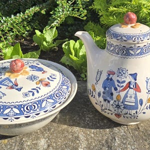 May include: A ceramic teapot and a matching lidded bowl featuring a cream-colored base with blue, red, and yellow folk-art style illustrations of figures and floral motifs. Both pieces have a rounded shape and a small, reddish-brown knob on the lid.