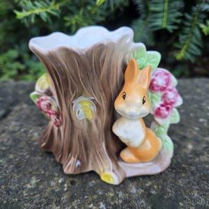 May include: A ceramic planter in the shape of a tree stump, with a seated orange and white bunny. The planter has a brown trunk with a white interior, and is decorated with pink flowers and green leaves. A decorative piece.