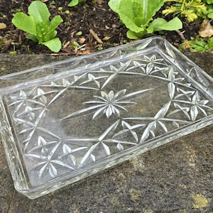 Puede incluir: Una bandeja rectangular de vidrio transparente con un diseño de estrella. La bandeja tiene un borde elevado y descansa sobre una superficie de piedra. El diseño de estrella está grabado en el vidrio, creando un efecto decorativo.