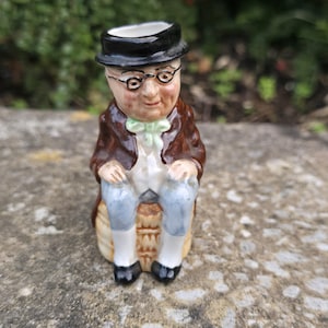 Può includere: Una statuetta in ceramica raffigurante una persona con cappello nero, occhiali tondi, cappotto marrone e papillon verde chiaro. La figura è seduta su una base cilindrica testurizzata color beige, indossando pantaloni azzurri e scarpe nere.