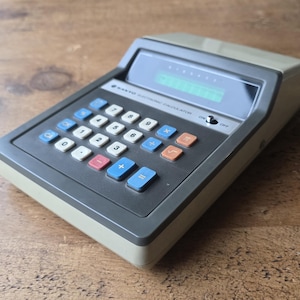 Könnte beinhalten: Vintage elektronischer Sanyo-Taschenrechner mit grauem und beigem Gehäuse. Der Rechner verfügt über einen Bildschirm und eine Tastatur mit blauen, weißen, roten und orangefarbenen Tasten. Der Text "SANYO ELECTRONIC CALCULATOR" ist auf dem Rechner aufgedruckt.