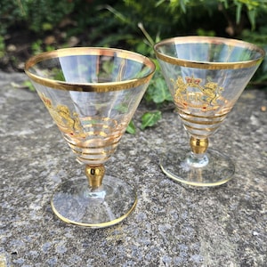 Op de afbeelding: Twee heldere glazen op voet met gouden randen en horizontale gouden banden. Elk glas toont een gouden embleem met een kroon, een leeuw, een eenhoorn en de tekst ER Coronation 1953. De glazen hebben een conische kelk en een gouden steel.