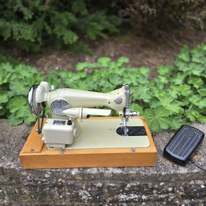 May include: A vintage off-white sewing machine with a wooden base and a black foot pedal. The machine has a chrome needle guard and a light beige body. The machine is on a stone surface with green foliage in the background.