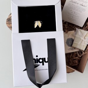 May include: A gold and silver leaf-shaped ring displayed in a white box with a black interior. The box is inside a white shopping bag with black handles. The bag has the word "niqu" printed on it. A gift box and a card are visible.