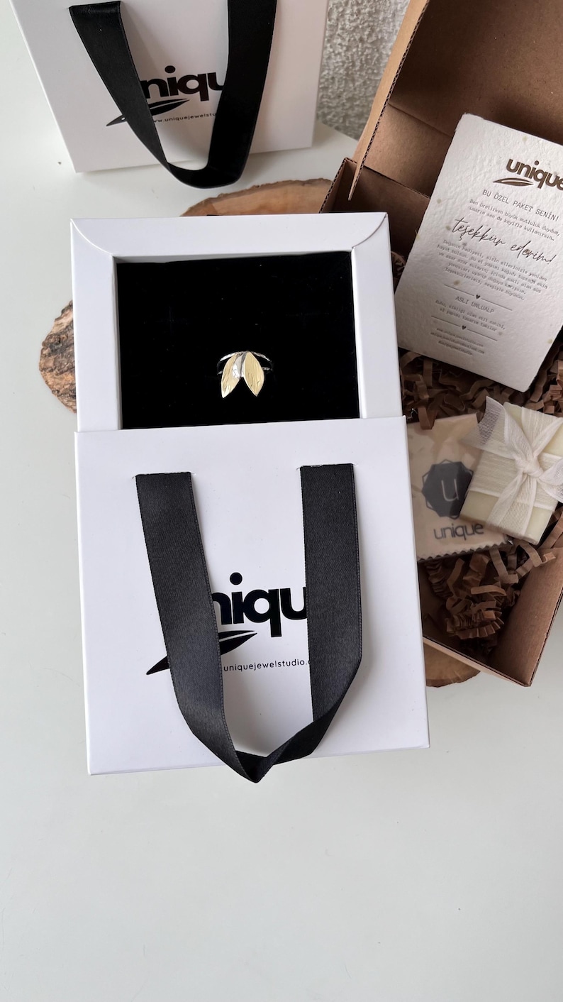 May include: A gold and silver leaf-shaped ring displayed in a white box with a black interior. The box is inside a white shopping bag with black handles. The bag has the word "niqu" printed on it. A gift box and a card are visible.