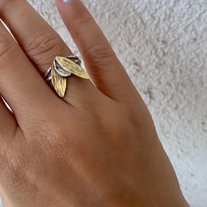 May include: A silver ring with two gold-colored leaf-shaped accents. The ring has a textured band and is worn on a finger. The background is a textured white wall.