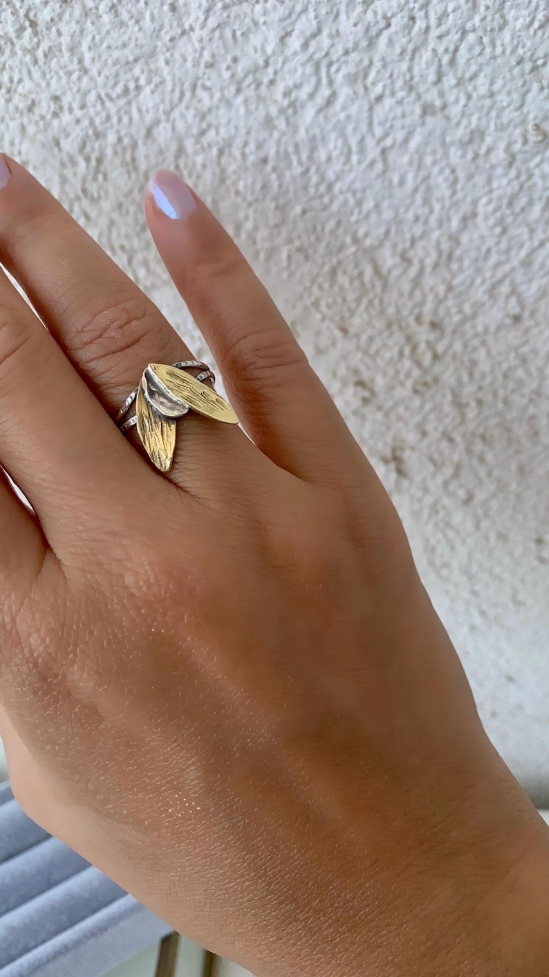 May include: A silver ring with two gold-colored leaf-shaped accents. The ring has a textured band and is worn on a finger. The background is a textured white wall.