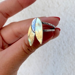 May include: A silver ring with two leaf-shaped gold and silver accents. The leaves have a textured surface and are attached to a silver band with etched details. The ring is a piece of jewelry.
