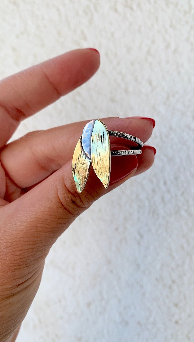 May include: A silver ring with two leaf-shaped gold and silver accents. The leaves have a textured surface and are attached to a silver band with etched details. The ring is a piece of jewelry.