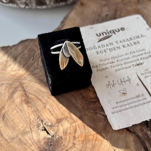 May include: A silver ring with two gold-colored leaf-shaped pendants, displayed in a black velvet box. The ring is on a wooden surface, with a paper card and a decorative stone in the background. The ring has a unique design.