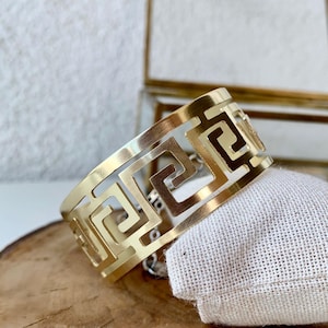 May include: Gold-toned metal bracelet with a geometric pattern. The bracelet is on a wooden surface.