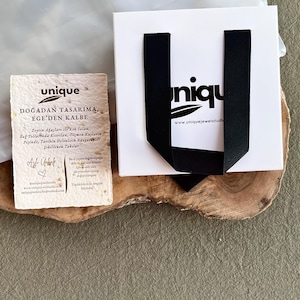 May include: A white gift bag with black ribbon handles and the word "unique" printed on it. A small card with text in Turkish and the word "unique" is next to the bag. The items are on a wooden surface.