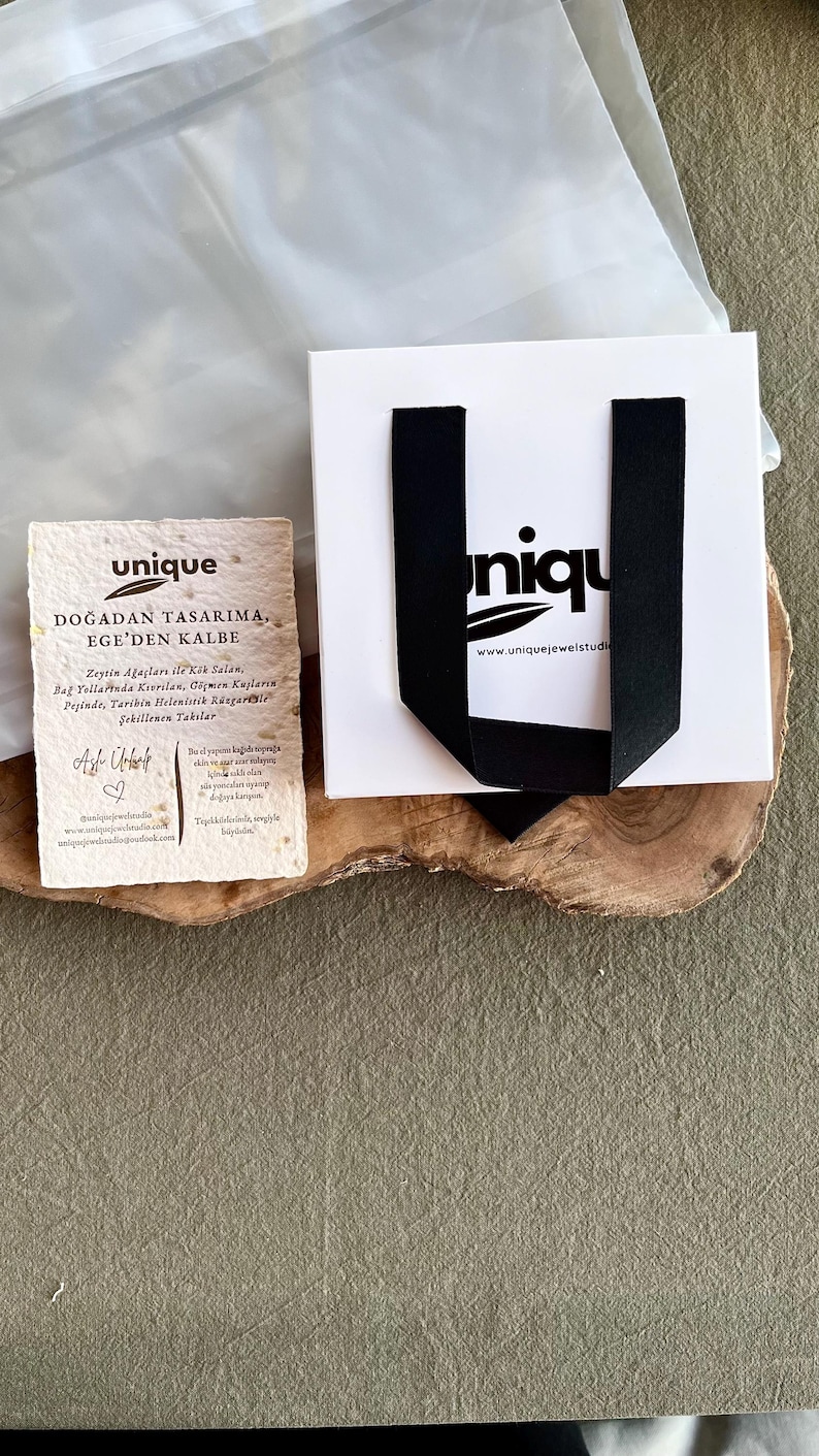 May include: A white gift bag with black ribbon handles and the word "unique" printed on it. A small card with text in Turkish and the word "unique" is next to the bag. The items are on a wooden surface.