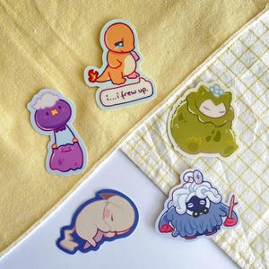 Op de afbeelding: Een verzameling kleurrijke stickers met stripfiguren. Eén sticker toont een oranje wezen met de tekst "i...frew up." Andere stickers tonen verschillende wezens in paars, groen en blauw. De stickers zijn gerangschikt op een geel en wit patroon.