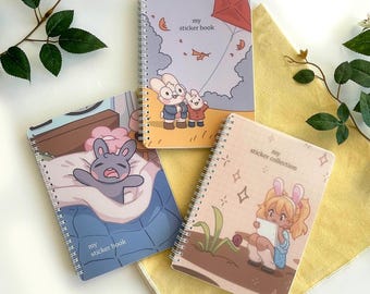 Puede incluir: Tres cuadernos con espiral con cubiertas ilustradas. Uno presenta un conejito en la cama, otro muestra conejitos con una cometa, y el tercero representa a una niña con orejas de conejo. Cada cuaderno tiene el texto "my sticker book" o "my sticker collection".