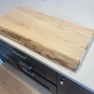 Può includere: Un tagliere rettangolare in legno con bordo naturale. Il legno di colore chiaro ha una venatura visibile e un bordo più scuro e contrastante. Il tagliere è posto su un piano di lavoro bianco sopra mobili scuri.