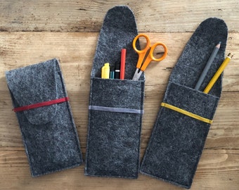 May include: Three gray felt pencil cases with colorful elastic bands and pockets. One case is closed with a red band, the others are open, displaying pens, pencils, and orange scissors. The cases are on a wooden surface.
