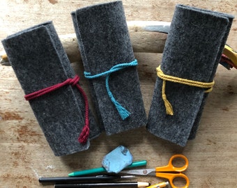 May include: Three gray felt pencil cases tied with red, teal, and yellow cords. A blue eraser, green and black pencils, a yellow pencil, and orange scissors are arranged below the cases. The items are on a wooden surface.