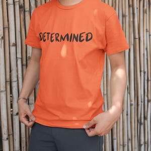 Puede incluir: Camiseta naranja con la palabra "DETERMINED" impresa en negro. La camiseta de manga corta tiene cuello redondo. La persona lleva pantalones grises. El fondo es una pared de bambú.