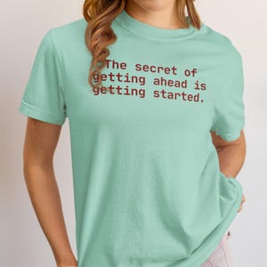 May include: A light teal t-shirt with the quote, "The secret of getting ahead is getting started," printed in a dark red font. The shirt has short sleeves and a relaxed fit.