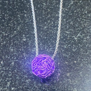 May include: A silver chain necklace featuring a woven purple sphere pendant. The pendant is a ball of intertwined metal strands, creating a textured, open design. The necklace is set against a dark, speckled background.