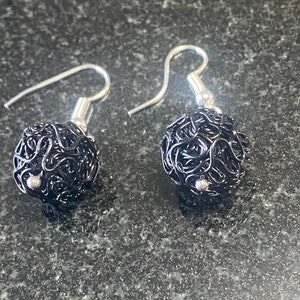 May include: A pair of black wire-wrapped ball earrings with silver-coloured ear wires. The earrings have a spherical shape and a textured, woven appearance. The earrings are displayed on a dark, speckled surface.
