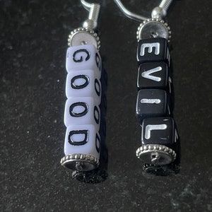 May include: A pair of handmade earrings. One earring spells out "GOOD" in white letter beads, the other spells "EVIL" in black letter beads. The earrings have silver-coloured hooks and decorative caps.