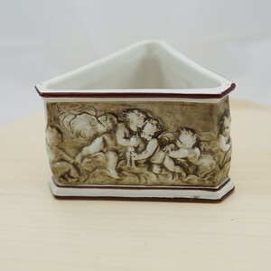 Può includere: Un vaso da fiori triangolare in ceramica con un design marrone e bianco con cherubini. Il vaso ha un bordo rosso.