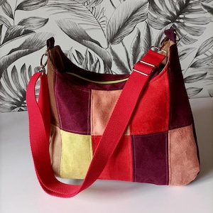 Op de afbeelding: Een patchwork schoudertas met een rode band. De tas is gemaakt van suède in verschillende kleuren, waaronder rood, bordeaux, geel en beige. De band is van felrode stof. De tas heeft een ritssluiting aan de bovenkant en een lichtgroene binnenkant.