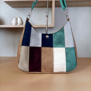 Peut inclure: Un sac à bandoulière de style hobo avec un motif patchwork composé de panneaux rectangulaires bleu marine, gris clair, bordeaux foncé, beige, blanc et sarcelle. Le sac comprend une fermeture à pression en métal argenté et une sangle réglable.