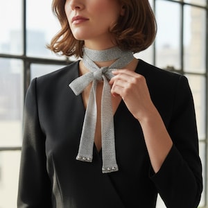 May include: A woman in a black jumpsuit, accessorised with a silver-grey necktie tied in a bow. The necktie features small silver details at the ends. A window and a wooden table with a plant and books are in the background.