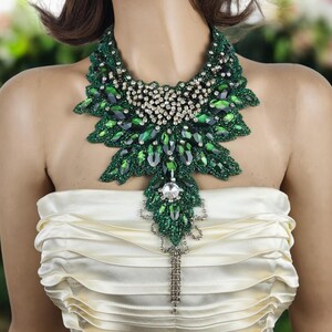 May include: A statement necklace featuring a large, ornate design. The necklace is primarily green with a leaf-like pattern, accented with clear crystals and a dangling chain. The base of the necklace appears to be beaded, and the overall style is elegant and eye-catching.