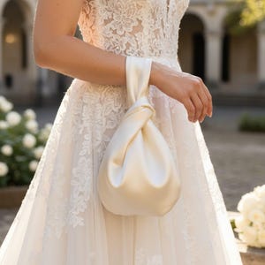 May include: Ivory satin bridal handbag with a knotted handle. The bag is held by a person wearing a white lace wedding dress. The background includes a blurred outdoor setting with white flowers and architectural details.