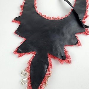 May include: A black leather necklace with a unique, asymmetrical shape. The edges are trimmed with red lace and beads. A black ribbon is attached for wearing. The necklace is a statement piece.