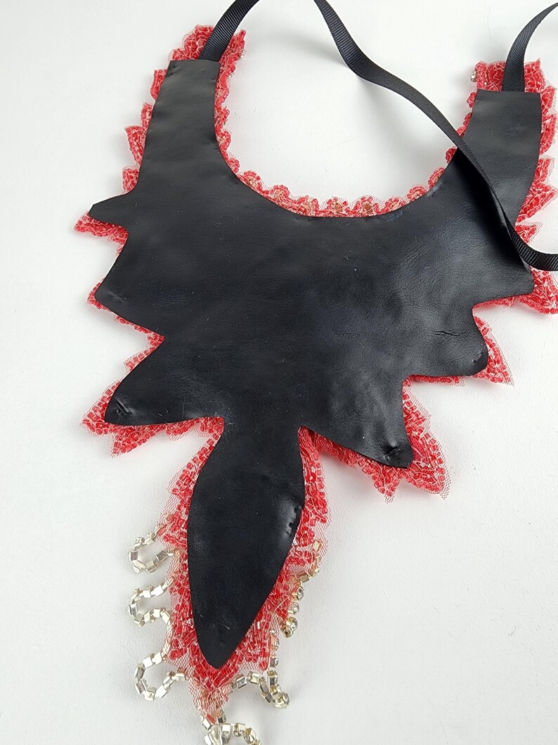 May include: A black leather necklace with a unique, asymmetrical shape. The edges are trimmed with red lace and beads. A black ribbon is attached for wearing. The necklace is a statement piece.