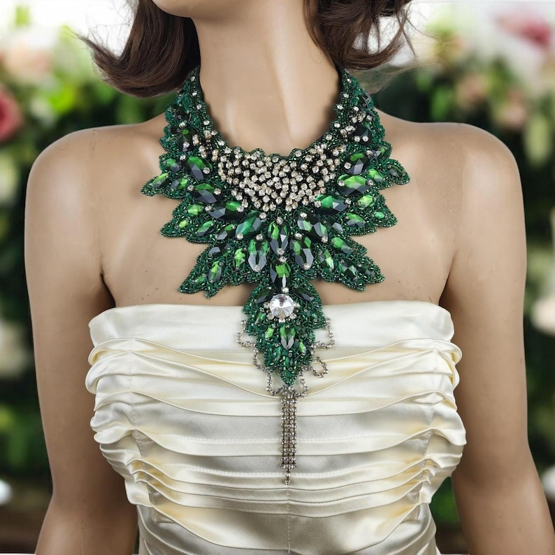 May include: A statement necklace featuring a large, ornate design. The necklace is primarily green with a leaf-like pattern, accented with clear crystals and a dangling chain. The base of the necklace appears to be beaded, and the overall style is elegant and eye-catching.