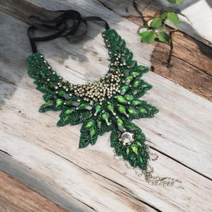 May include: A green beaded statement necklace with a leaf-like design. The necklace features emerald-green and clear crystal accents, with a central cluster of gold-toned beads. A black ribbon secures the necklace.