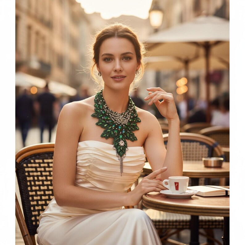 May include: A woman wearing a cream-colored strapless dress and a statement necklace with green and silver accents. The necklace features a leaf-like design. She is seated at a cafe table with a cup of coffee.