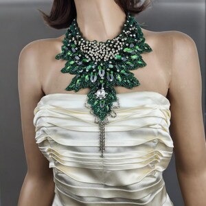 May include: A striking green and silver statement necklace. The necklace features large emerald-colored stones and smaller clear crystals, arranged in a leaf-like pattern. A long, dangling silver pendant hangs from the center. The necklace is displayed on a mannequin wearing a cream-colored strapless dress.