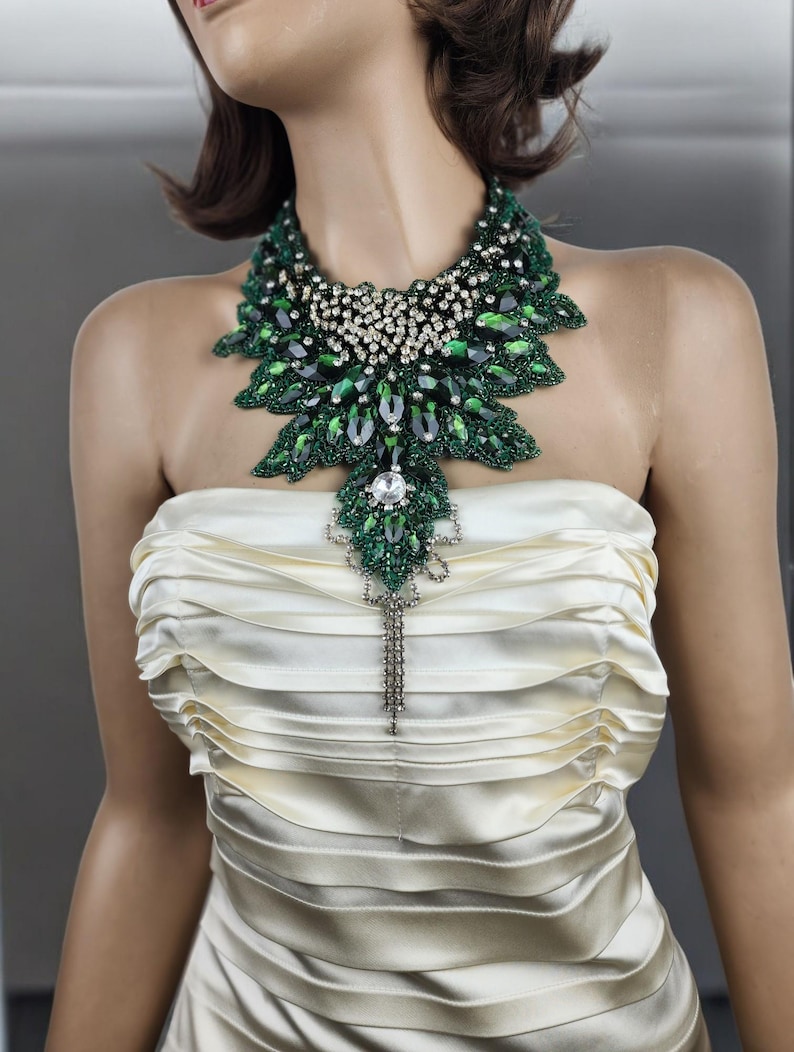May include: A striking green and silver statement necklace. The necklace features large emerald-colored stones and smaller clear crystals, arranged in a leaf-like pattern. A long, dangling silver pendant hangs from the center. The necklace is displayed on a mannequin wearing a cream-colored strapless dress.