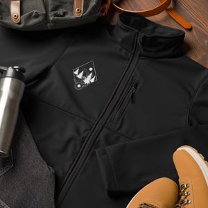 Può includere: Giacca softshell nera con un designo ricamato bianco di una scena di foresta con alberi e una luna. La giacca ha una chiusura a zip e due tasche.