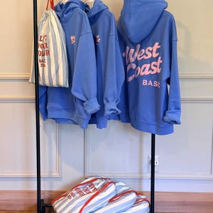 May include: Three periwinkle blue hooded sweatshirts with pink text reading West Coast BASIC on the back, hanging on a black metal rack. A white and blue striped drawstring bag with red text is hanging on the left, with two more bags on the bottom shelf.