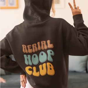 May include: A brown hoodie with the text "AERIAL HOOP CLUB" printed on the back in a retro, colourful font.