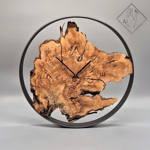 Könnte beinhalten: Eine runde Wanduhr mit einem Zifferblatt aus Naturholz und einem dunklen Metallrahmen. Das Zifferblatt der Uhr zeigt ein einzigartiges Holzmaserungsmuster, schwarze Zeiger und die römische Ziffer III. Die Uhr hat eine rustikale, organische Ästhetik.